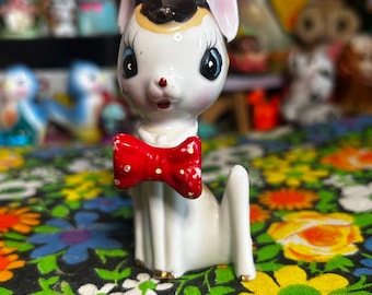 May include: A vintage ceramic figurine of a white dog with a red bow tie. The dog has black ears, a black and tan head, and large blue eyes. The figurine is on a floral patterned fabric.