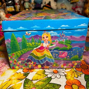 Puede incluir: Una caja decorativa con una vibrante ilustraci&oacute;n de una princesa con un vestido arco&iacute;ris, un castillo y un cisne sobre un fondo azul. La caja est&aacute; adornada con dise&ntilde;os florales y paisaj&iacute;sticos, creando una est&eacute;tica caprichosa y colorida.