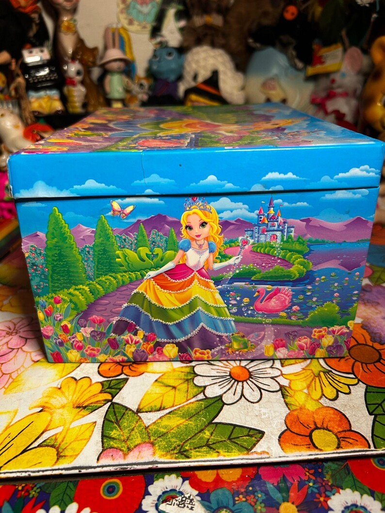 Puede incluir: Una caja decorativa con una vibrante ilustraci&oacute;n de una princesa con un vestido arco&iacute;ris, un castillo y un cisne sobre un fondo azul. La caja est&aacute; adornada con dise&ntilde;os florales y paisaj&iacute;sticos, creando una est&eacute;tica caprichosa y colorida.