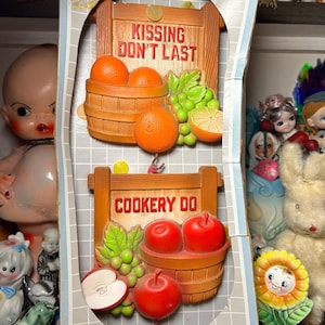 May include: Vintage kitchen hot pad holder with a wood frame and fruit design. The top sign reads "Kissing Don't Last" with oranges and grapes. The bottom sign reads "Cookery Do" with apples and grapes.