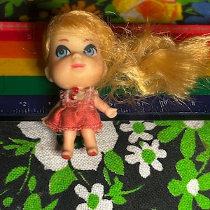 May include: A vintage doll with blonde hair and blue eyes, wearing a red dress. The doll is small, with painted facial features. The background includes a rainbow-colored ruler and a floral patterned fabric.