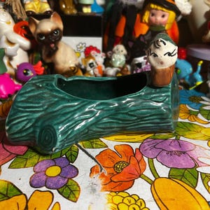 May include: A vintage, green ceramic planter shaped like a log, with a small figurine on one end. The planter has a textured surface and a dark interior. The background includes various figurines and a floral patterned surface.