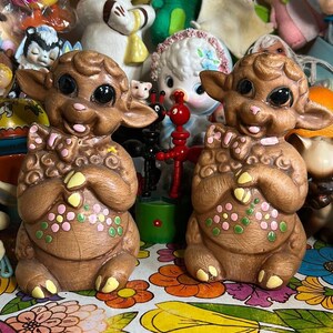 May include: Two brown ceramic lamb figurines with large black eyes and pink and yellow floral accents. The lambs are holding their front paws together in front of their chests. The figurines are sitting on a colorful floral patterned surface.