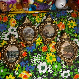 May include: Four vintage oval framed landscape paintings with ornate gold frames. The paintings depict scenes of trees, water, and buildings. The frames are arranged on a floral patterned fabric with a variety of colorful flowers.