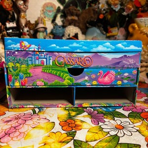 Puede incluir: Una colorida caja de almacenamiento Princess Cassara con un caj&oacute;n y dos compartimentos. La caja presenta un paisaje caprichoso con un castillo, un cisne y el texto "Princess Cassara". La caja est&aacute; colocada sobre una superficie con estampado floral.