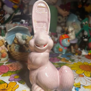 Puede incluir: Figura vintage de conejo de Pascua de cerámica rosa con una oreja alta y rectangular y detalles rojos. El conejo está sentado y está rodeado de otros juguetes y objetos de colección vintage.