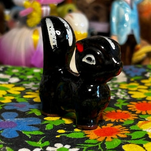 May include: A glossy black ceramic skunk figurine with a white stripe down its back and tail, and a red spot on its head. The skunk is standing on a floral patterned surface, with other figurines in the background.