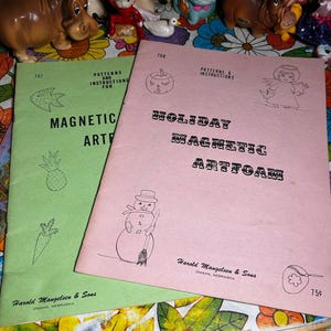 May include: Two vintage craft pattern books, one green and one pink, with the titles "Magnetic Art" and "Holiday Magnetic Artfoam." The covers feature illustrations of a pineapple, carrot, pumpkin, angel, and snowman. The books are from Harold Mangelsen & Sons, Omaha, Nebraska.