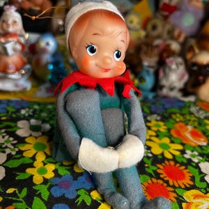 May include: A vintage doll with a painted face, red hair, and a gray and blue outfit. The doll has a red collar and a gray hat with a white brim. The doll is sitting on a floral patterned fabric.