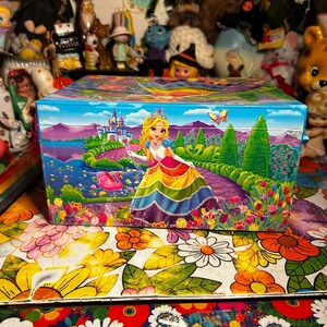 Puede incluir: Una caja rectangular colorida con una ilustraci&oacute;n de princesa. La caja presenta una princesa con un vestido arco&iacute;ris, un castillo y una mariposa. El fondo muestra un lago y un jard&iacute;n. La caja est&aacute; sobre una superficie con estampado floral.