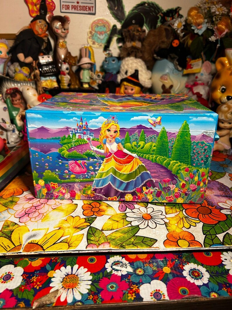 Puede incluir: Una caja rectangular colorida con una ilustraci&oacute;n de princesa. La caja presenta una princesa con un vestido arco&iacute;ris, un castillo y una mariposa. El fondo muestra un lago y un jard&iacute;n. La caja est&aacute; sobre una superficie con estampado floral.