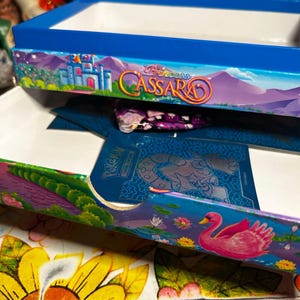 Puede incluir: Una caja azul con el texto "Princess Cassara" y una ilustraci&oacute;n de un castillo. La caja tiene un interior blanco y est&aacute; decorada con coloridos dise&ntilde;os florales y paisaj&iacute;sticos. Un cisne rosa aparece en el lateral.