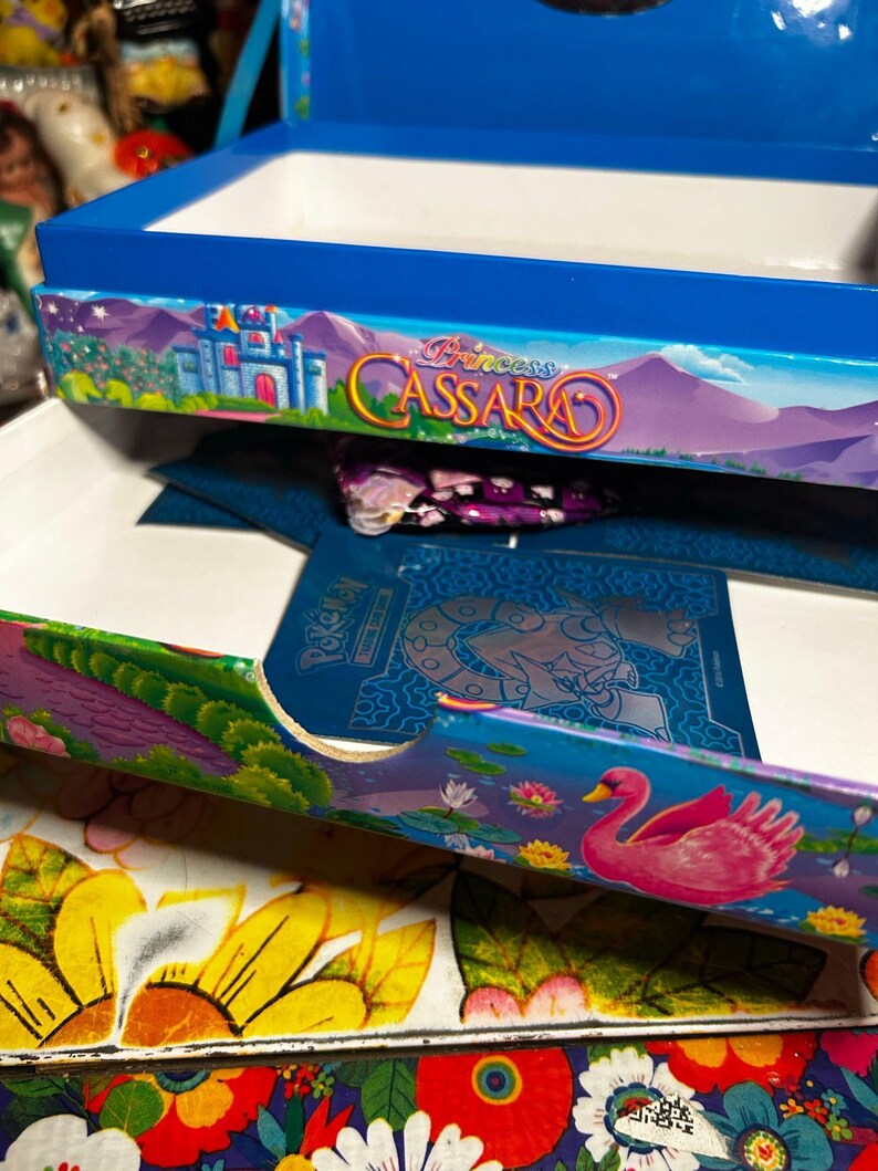 Puede incluir: Una caja azul con el texto "Princess Cassara" y una ilustraci&oacute;n de un castillo. La caja tiene un interior blanco y est&aacute; decorada con coloridos dise&ntilde;os florales y paisaj&iacute;sticos. Un cisne rosa aparece en el lateral.