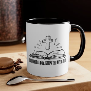 Puede incluir: Taza de cerámica blanca con interior y asa negros. La taza presenta una ilustración en blanco y negro de una cruz sobre un libro abierto, con el texto "A PROVERB A DAY, KEEPS THE DEVIL AWAY".