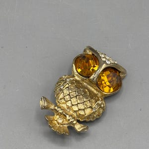Pode incluir: Broche de cor dourada em forma de coruja com grandes olhos de strass âmbar e pequenos strass transparentes. O corpo da coruja tem um design texturizado e padronizado. O broche é um acessório de estilo vintage.