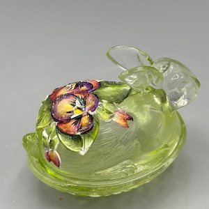 May include: A light green glass bunny figurine with a detailed floral design. The bunny has clear glass ears and is decorated with a colorful pansy and leaves. The flowers have purple, yellow, and orange hues.