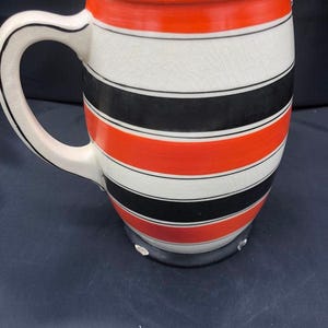 Może przedstawiać: Ceramiczny dzbanek z białym wnętrzem i uchwytem. Zewnętrzna strona posiada poziome paski w kolorze czerwonym, czarnym i białym. Górna krawędź jest czerwona, a uchwyt biały z czarnym paskiem.