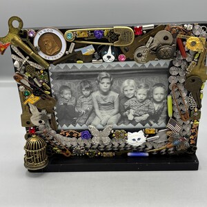 May include: A rectangular black picture frame decorated with an assortment of metal keys, safety pins, a birdcage, a butterfly, a cat face, and a coin inscribed with In God We Trust. The frame holds a black and white photograph of six children.