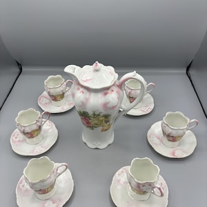 Może przedstawiać: Biały porcelanowy zestaw do kawy składający się z wysokiego dzbanka i pięciu pasujących filiżanek ze spodkami. Każdy element ozdobiony jest różowymi akcentami kwiatowymi i motywami żółtych róż, wykończony falistymi brzegami i delikatnym różowym odcieniem.