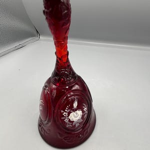 Puede incluir: Una campana de cristal rojo rubí con un mango largo. La campana presenta diseños florales blancos y es probablemente un artículo decorativo. La campana está hecha de vidrio transparente y tiene un acabado brillante.
