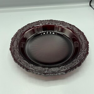 May include: A deep red glass plate featuring a smooth, circular centre and a wide, decorative rim. The rim is adorned with a repeating pattern of embossed diamond shapes and small floral motifs, finished with a scalloped, wavy edge.