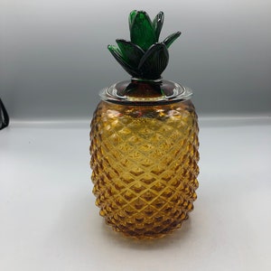 May include: A glass pineapple-shaped jar with a green glass pineapple top. The jar is amber in colour and has a textured surface.