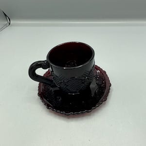 Puede incluir: Un juego de taza y platillo de vidrio rojo rubí. La taza tiene un asa y se asienta sobre un platillo a juego. Ambas piezas presentan un patrón en relieve detallado. El juego está hecho de vidrio rojo oscuro.
