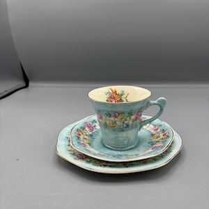 Könnte beinhalten: Ein Vintage-Tee-Set mit Tasse, Untertasse und Kuchenteller. Das Set hat einen hellblauen Hintergrund mit einem Blumenmuster in Rosa, Gelb und Blau. Die Ränder sind goldfarben verziert. Die Tasse hat einen Henkel und ein passendes Blumenmuster im Inneren.