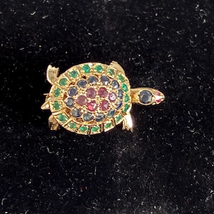 Op de afbeelding: Een goudkleurige schildpadbroche met een schild versierd met een mozaïek van kleurrijke edelstenen. De schildpad heeft groene, blauwe en rode stenen. De kop van de schildpad heeft een rode edelsteen oog.