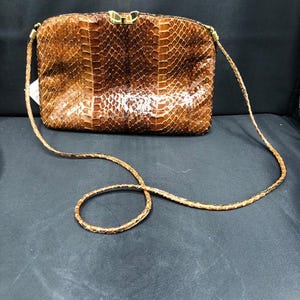 May include: A brown snakeskin-effect handbag with a gold clasp and a long, braided strap. The rectangular bag has a glossy finish and a textured surface, suggesting a vintage or luxury item.