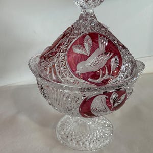 May include: A clear, cut-glass candy dish with a lid, featuring a ruby red interior and bird and leaf designs. The dish sits on a pedestal base and has a decorative handle on the lid. The glass is intricately cut with geometric patterns.