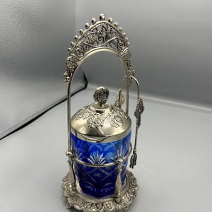 Puede incluir: Un tarro de cristal azul cobalto tallado con marco, tapa y asa de metal plateado. La tapa presenta racimos de uvas en relieve, y el marco incluye un asa arqueada ornamentada y una cuchara a juego colgada en el lateral del conjunto.