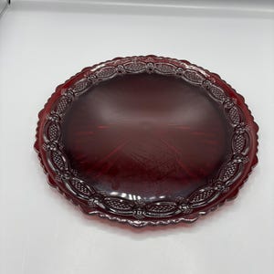 Puede incluir: Una fuente ovalada de vidrio rojo rubí. La fuente tiene un patrón detallado y en relieve alrededor del borde, con un borde festoneado. El centro de la fuente tiene una superficie lisa y reflectante.