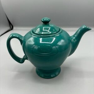Puede incluir: Una tetera de cerámica de color verde azulado con cuerpo redondeado y tapa. La tetera tiene asa y pico.