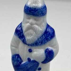 Op de afbeelding: Een klein, wit, getextureerd beeldje van een figuur met een baard, gekleed in een blauwe muts, blauwe kraag en blauwe wanten. Het figuur houdt een blauwe tas voor zich, met blauwe knopen zichtbaar op de witte jas. Het oppervlak is korrelig.