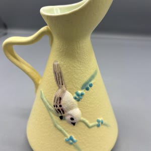 May include: A pale yellow ceramic pitcher with a curved handle and spout. A detailed bird and branch design is painted on the front. The bird is white and brown, and the branch has blue accents.
