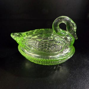 Puede incluir: Un joyero de vidrio verde en forma de cisne. El plato es de color verde vibrante y presenta una superficie texturizada en el cuerpo del cisne y un borde detallado en la base. El cuello del cisne forma un asa. Un artículo decorativo.