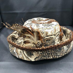 May include: A decorative hat with a snakeskin pattern in shades of brown, tan, and cream. The hat features a brown trim and is adorned with several brown and tan feathers. The hat has a wide brim and a rounded crown.
