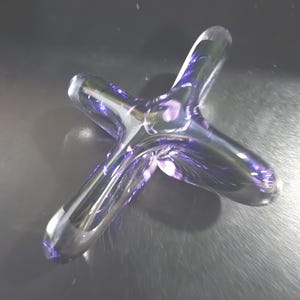 May include: A clear glass sculpture in a star-like shape with three elongated arms. The glass has a purple, iridescent sheen, with streaks of color throughout. The object is set against a dark background.