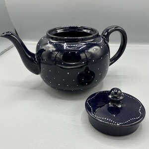 May include: A black ceramic teapot with a matching lid. The teapot has a rounded body, a curved spout, and a handle. It is decorated with small white polka dots and a white dotted border around the rim and lid.