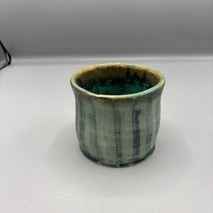 May include: A ceramic cup with a textured, matte finish featuring vertical dark blue-green stripes over a muted gray base. The interior of the cup is a vibrant, deep turquoise, while the rim shows a warm, earthy brown tone with a rough, uneven edge.
