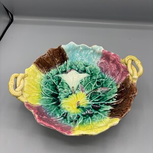 Op de afbeelding: Een kleurrijke keramische serveerschaal in de vorm van een koolblad. De schaal heeft een verscheidenheid aan kleuren, waaronder groen, geel, bruin, blauwgroen en roze. Het heeft twee gele handvatten in touwstijl en is bedoeld voor het serveren van voedsel.