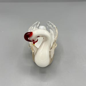 Op de afbeelding: Een wit glazen zwanenbeeldje met een rode en gele snavel. De zwaan zit in een gekrulde houding met uitgespreide vleugels.