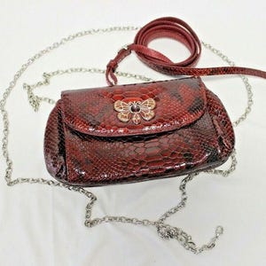 Könnte beinhalten: Burgunderfarbene Umhängetasche mit Schlangenmuster, silberner Kette und passendem burgunderfarbenem Lederriemen. Die Tasche hat einen dekorativen Schmetterlingsverschluss mit Strasssteinen.