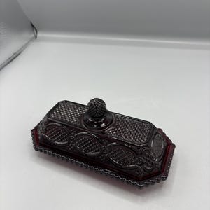Puede incluir: Una mantequera rectangular de vidrio rojo rubí con una tapa texturizada con diseño de diamantes y una base decorativa de forma ovalada. La tapa presenta un pequeño pomo redondo. El plato tiene un borde festoneado.