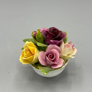 Puede incluir: Un arreglo floral de porcelana con un ramo de rosas amarillas, rosas y moradas en un tazón blanco. Las rosas están pintadas a mano y tienen un aspecto realista.