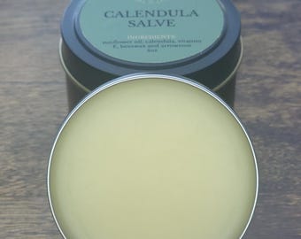 May include: A round, open tin of Calendula Salve, with a light yellow balm visible. The tin has a dark green lid with white text that reads "Calendula Salve" and "Herbcore". The ingredients are listed as sunflower oil, calendula, vitamin E, beeswax, and arrowroot, 4oz.