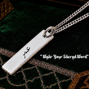 Op de afbeelding: Zilveren ketting met een rechthoekige hanger met Arabische kalligrafie. De hanger, gegraveerd met een naam, hangt aan een ketting. De tekst "Wear Your Sacred Word" is ook zichtbaar. Onderdeel van de Arabic Calligraphy Collection.