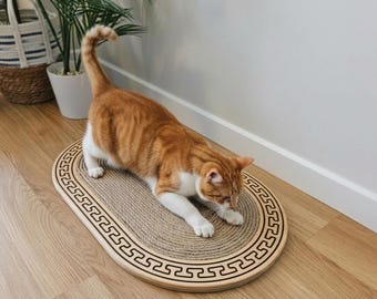 May include: An oval cat scratching pad featuring a light-colored wooden frame with a black Greek key pattern border and a textured, coiled sisal rope center, currently being used by an orange and white tabby cat.