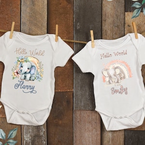 May include: Two white baby onesies with short sleeves. The onesies feature a rainbow and an elephant with floral accents. One onesie says "Hello World I am Harry" and the other says "Hello World I am Emily".